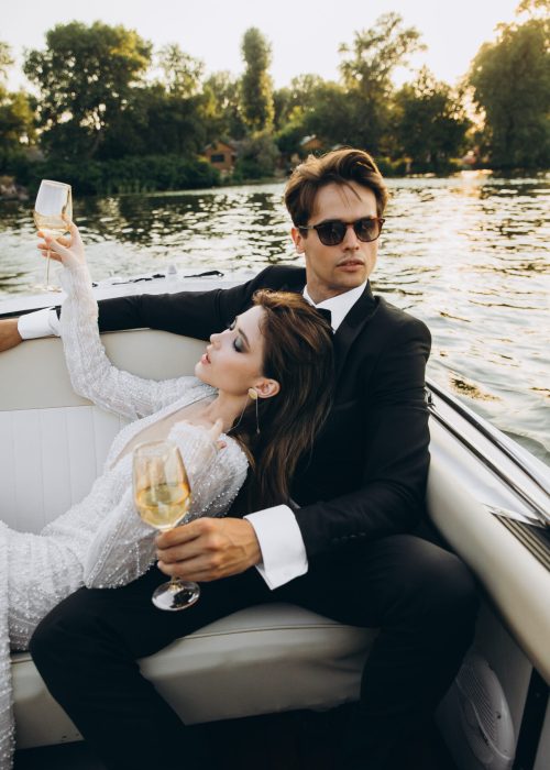 Groom in a black suit and sunglasses hugs bride in wedding dress on a yacht on a sunny day. Couple drinking champagne and having fun